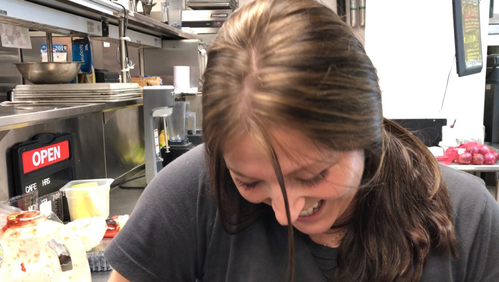 Michelle Hollingsworth is in the foreground with the kitchen of the Combine Cafe in the background. She is looking down while smiling and laughing. 