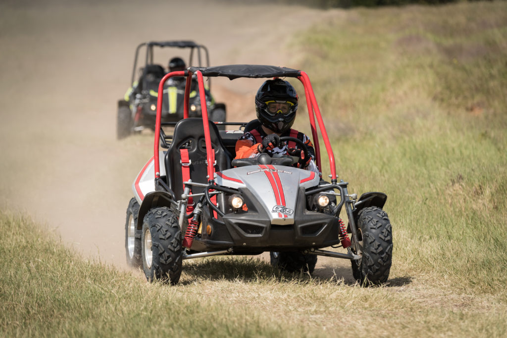 A silver Hammerhead GTS 150, with red accents, is turning a corner on a grassy lane. It is closely followed by a black GTS 150 with green accents. Both go karts have dirt clouds following them as they continue racing in the grass.